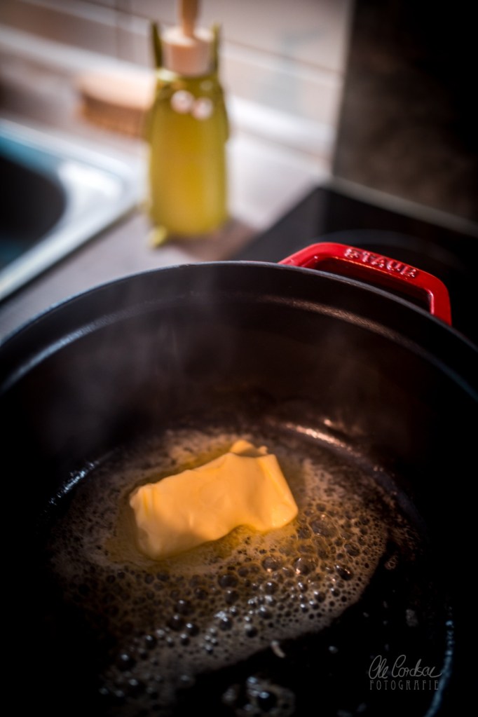 Ein Stück Butter schmilzt in einer Pfanne mit blubberndem Fett, im Hintergrund eine Küchenzeile.